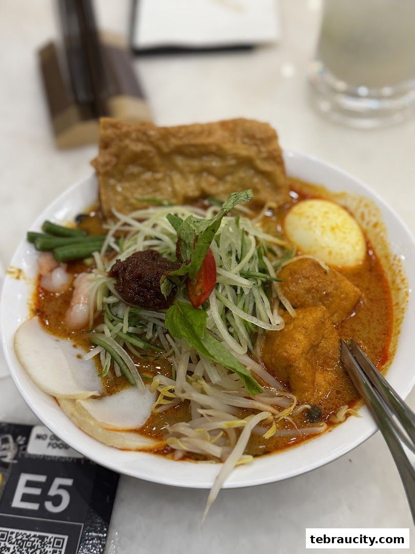 A close-up of a delicious bowl of Laksa with various toppings and a drink in the background.