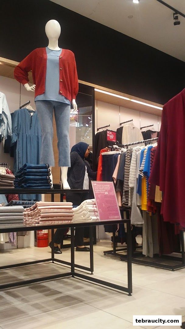 A mannequin displays a red cardigan and blue top, with clothing racks and folded garments in a store.