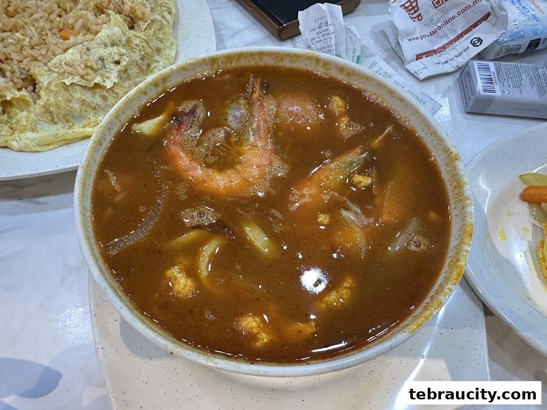 A close-up of a steaming bowl of curry soup with prawns and other ingredients, next to fried rice.