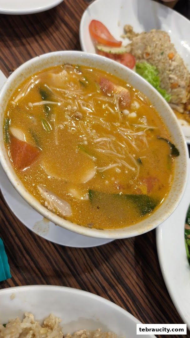 A close-up of a steaming bowl of Tom Yum soup with visible ingredients and a side of fried rice.