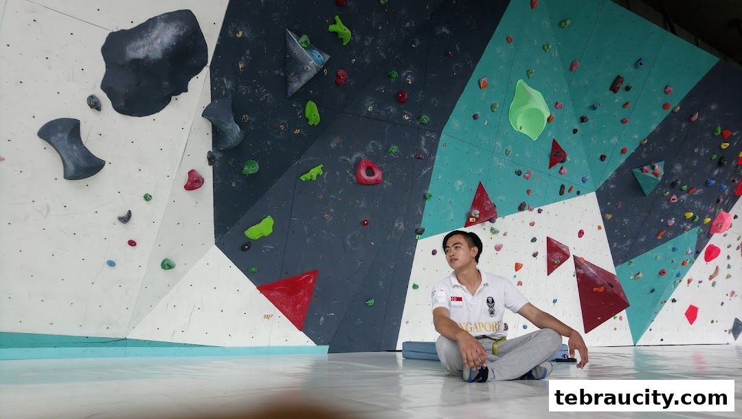 A young man sits on the floor in front of a colorful indoor climbing wall.