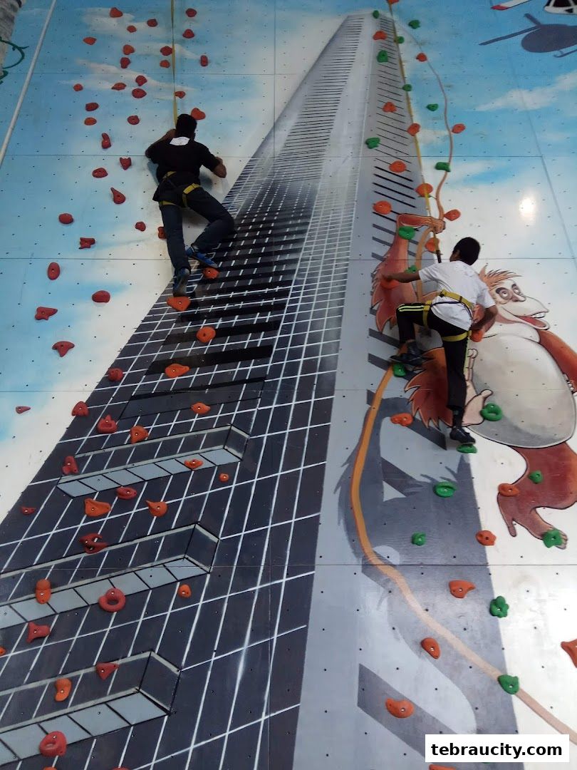 Two people are climbing a colorful indoor rock climbing wall with a cityscape design.