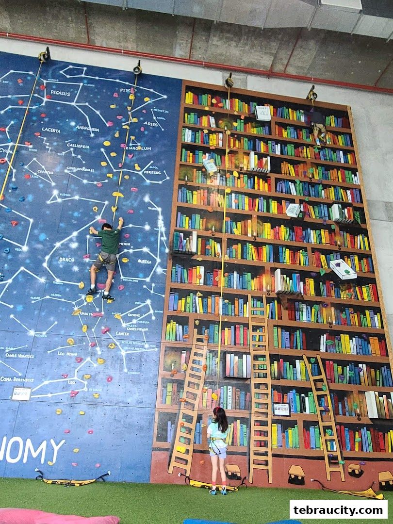 Two children are climbing a colorful indoor climbing wall designed like a bookshelf and a constellation map.