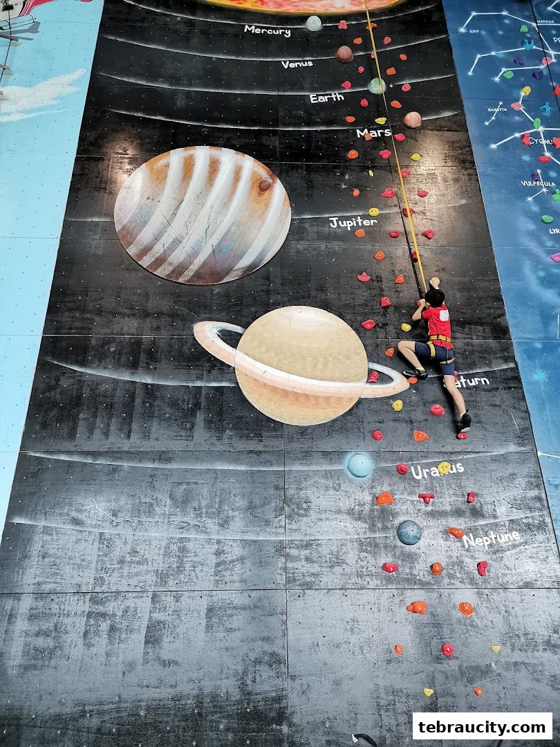 A young climber ascends a themed climbing wall depicting the solar system.