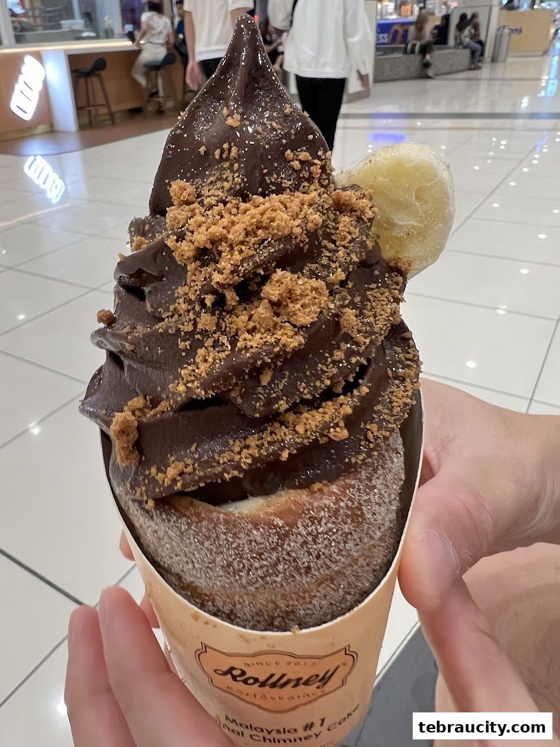 A close-up of a chocolate chimney cake with banana and cookie crumble toppings.