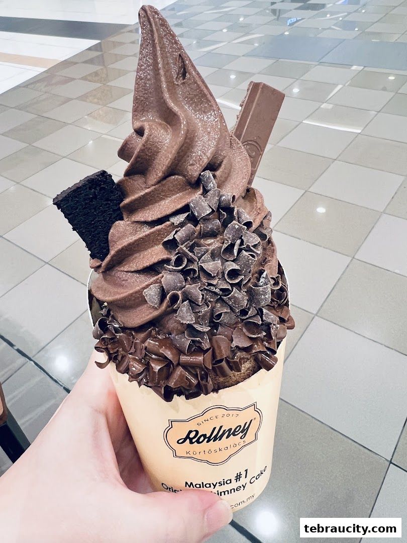 A hand holds a chocolate soft-serve ice cream cone topped with chocolate shavings and a Kit Kat.