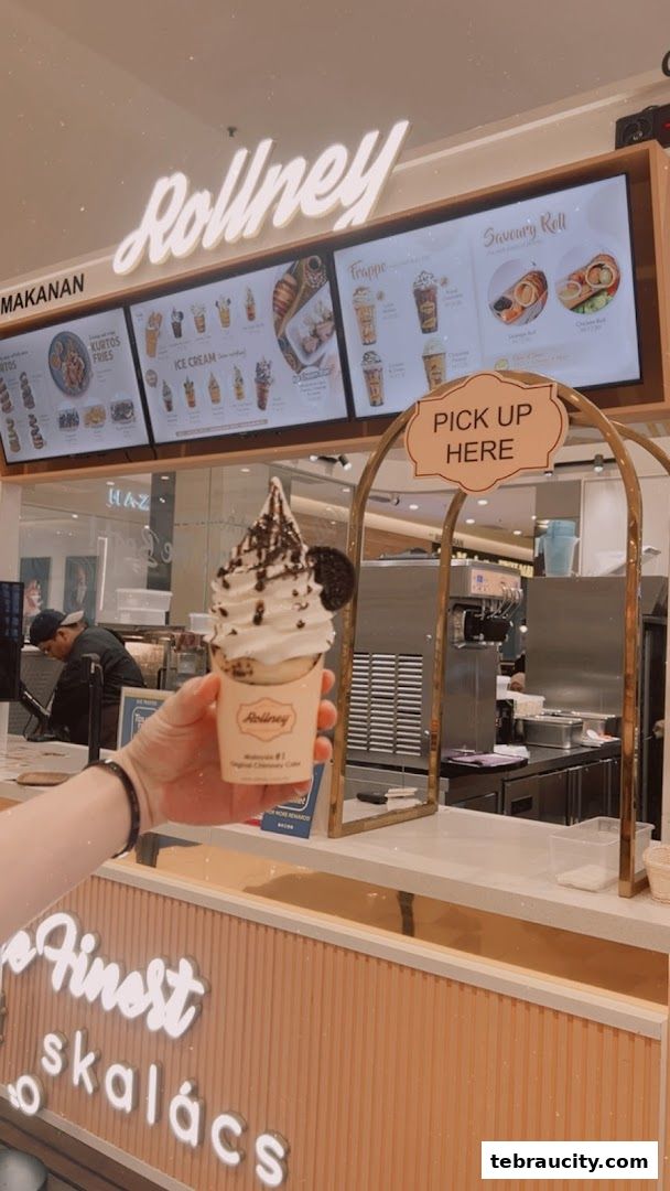 A hand holds a soft-serve ice cream cone in front of the Rollney shop counter.