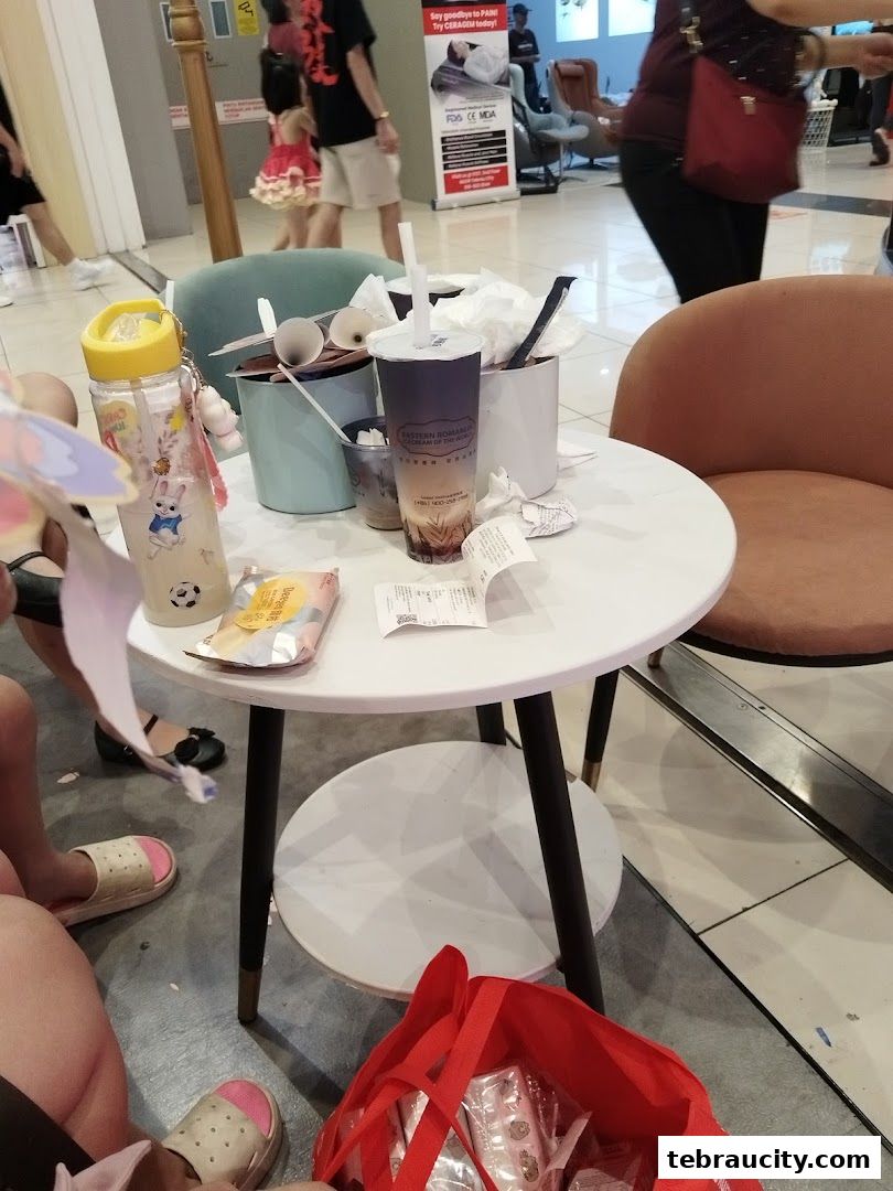 A table with drinks and trash, with people sitting around it in a mall.