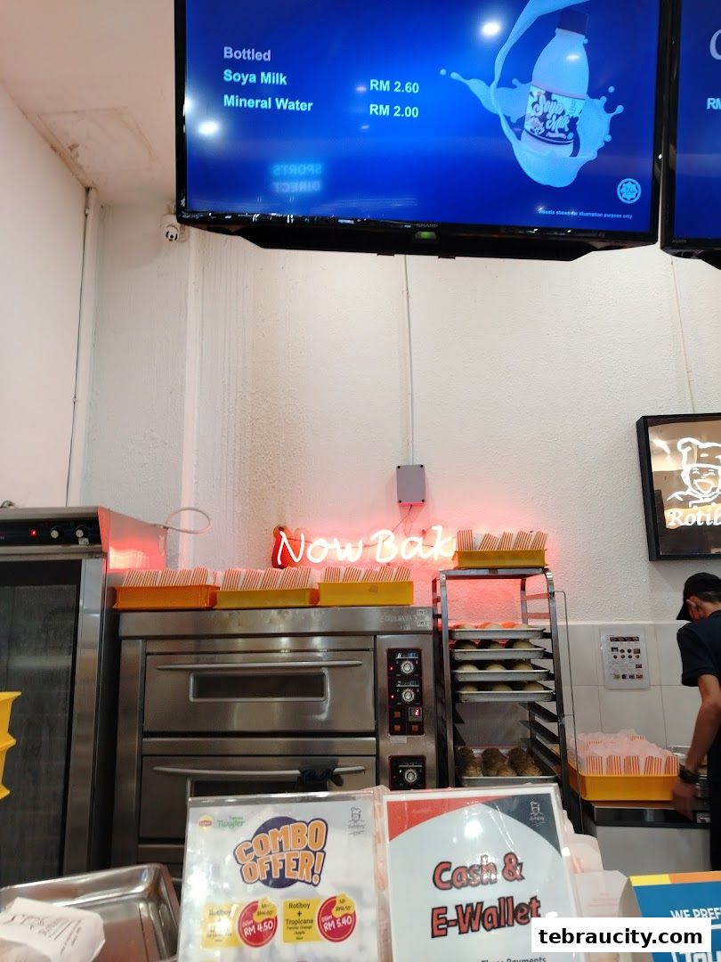 Interior view of a Rotiboy bakery with ovens, trays of baked goods, and a digital menu.
