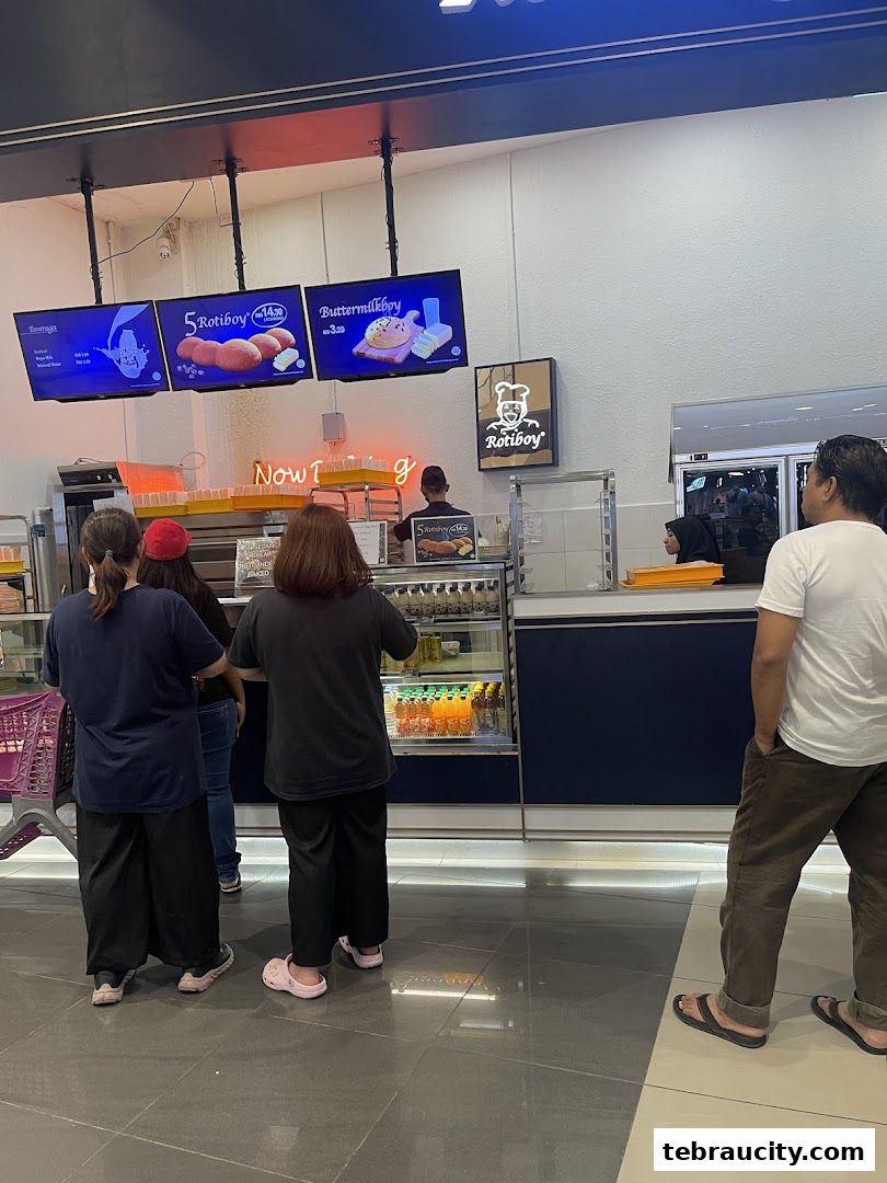 Customers queue at a Roti Boy counter with digital menus displaying products and prices.