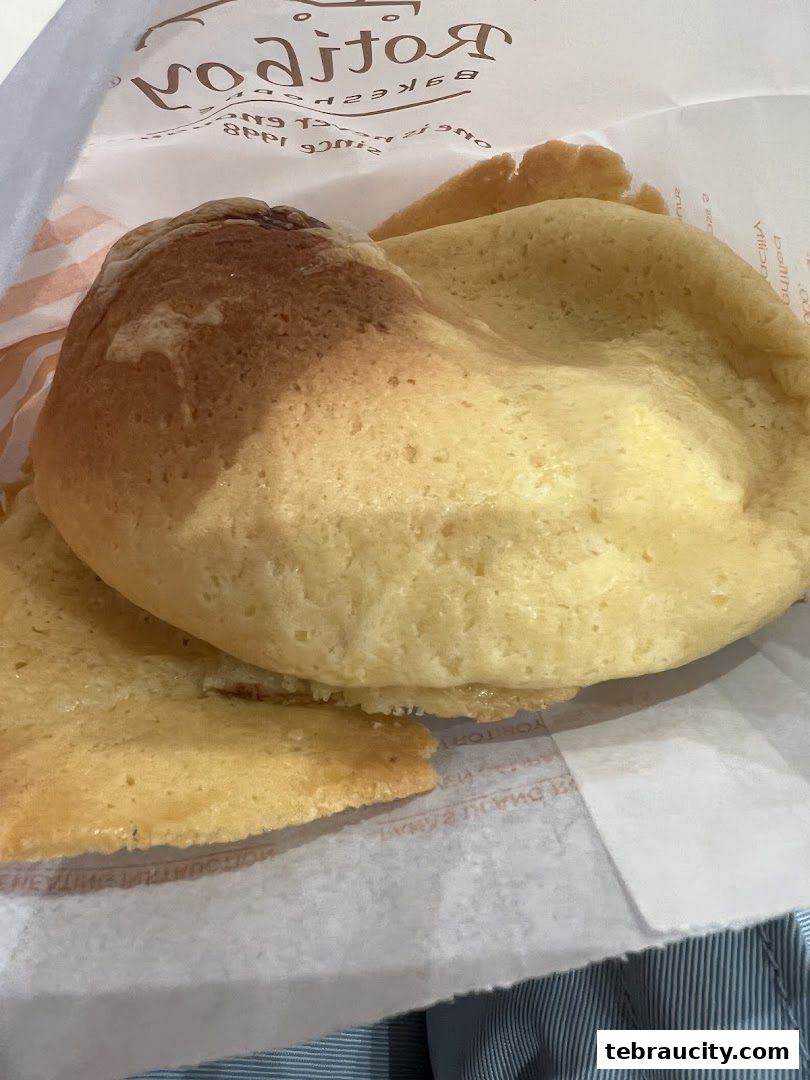 A close-up of a golden-brown baked bun with a crispy topping, presented in a paper bag.