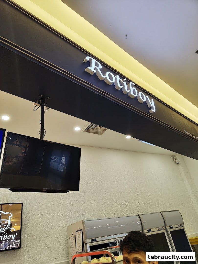 The Rotiboy shop sign is illuminated above a display of baked goods and a television screen.