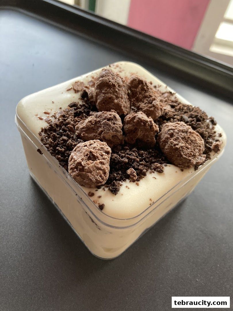 A close-up of a dessert in a clear plastic container, topped with chocolate pieces and crumbs.