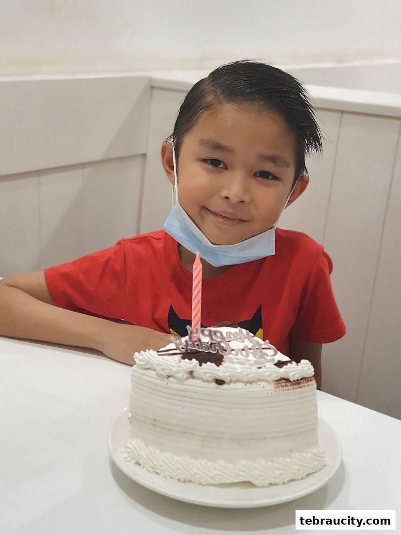A young boy in a red shirt sits at a table with a birthday cake and a candle.