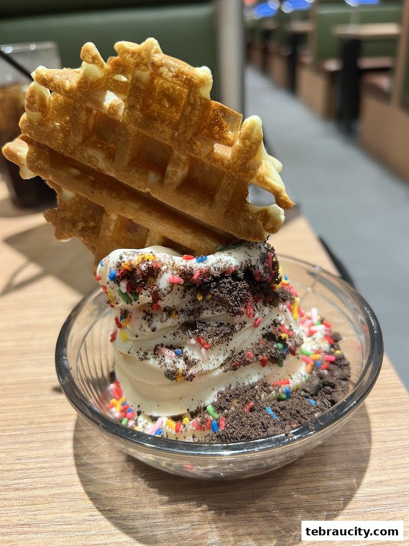 A close-up of a soft-serve ice cream topped with sprinkles and cookie crumbles, served with a waffle.