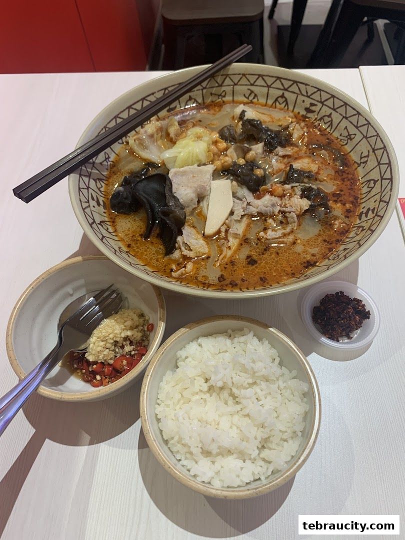 A bowl of spicy mala noodles with pork, vegetables, and a side of rice and chili garlic.