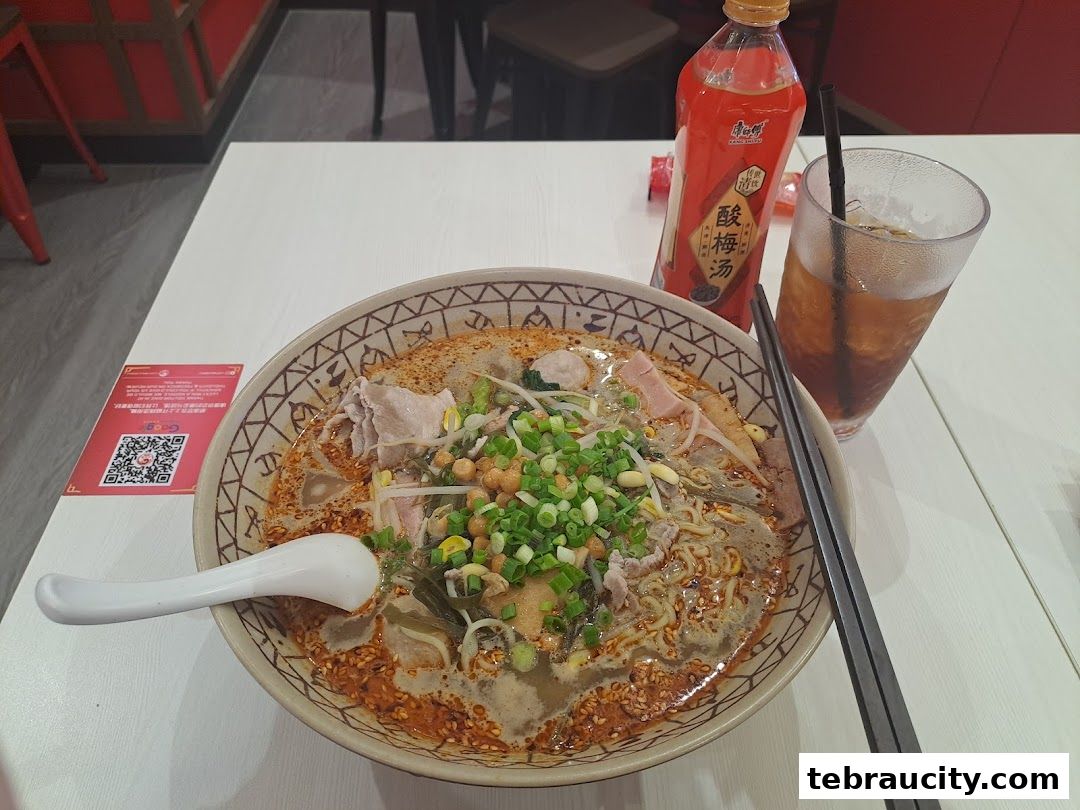 A bowl of mala noodles with a bottle of plum juice and a glass of iced tea.