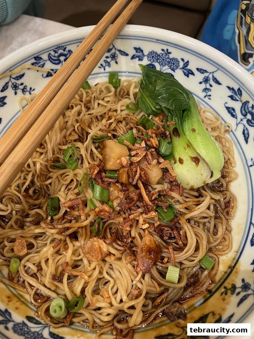 A close-up of a bowl of noodles with chopsticks, garnished with green onions and fried shallots.