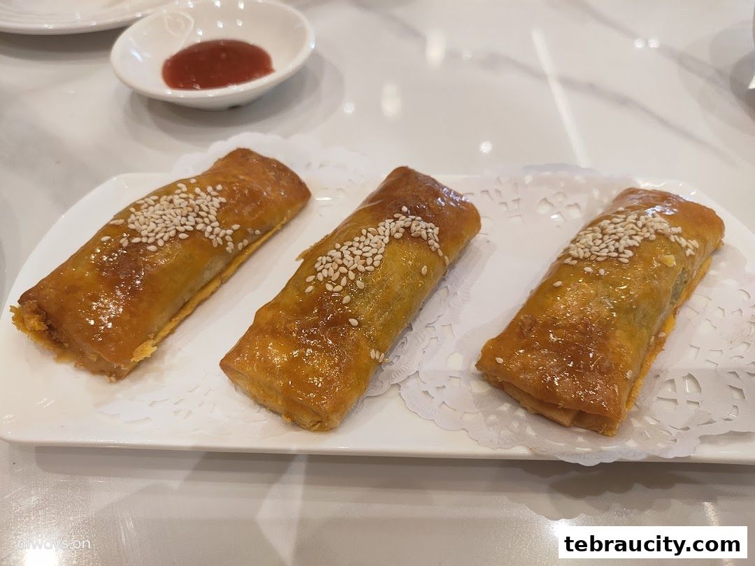 Three golden-brown spring rolls topped with sesame seeds on a white plate with dipping sauce.