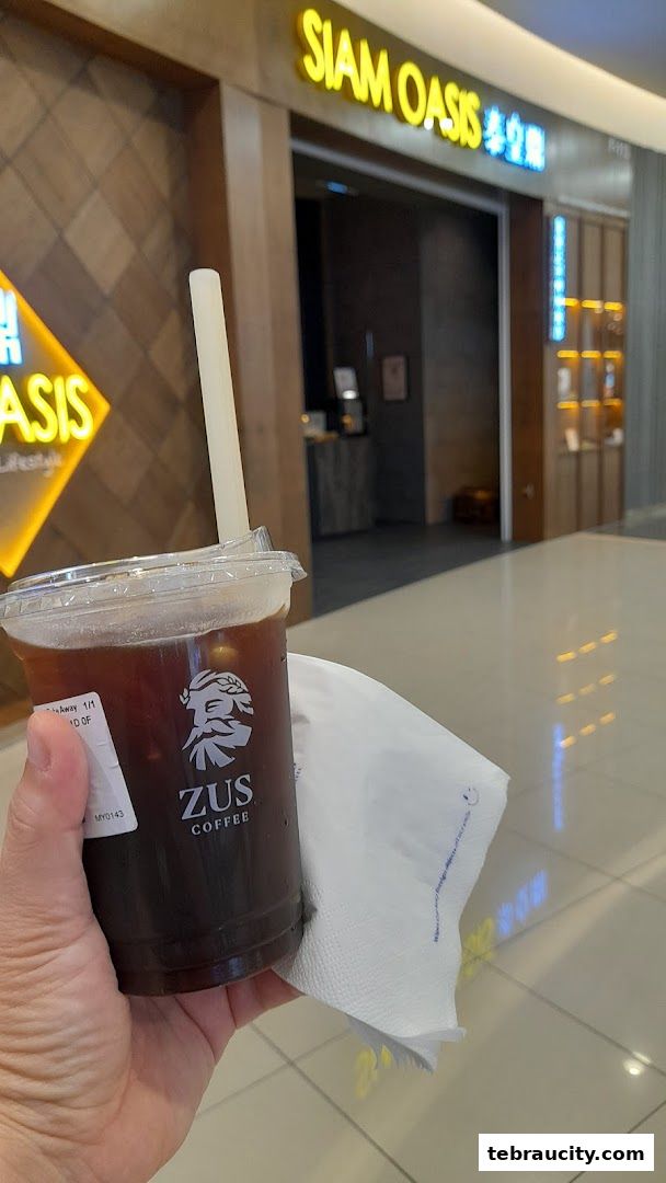 A hand holds a ZUS Coffee drink in front of the Siam Oasis store.