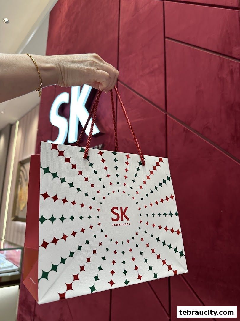 A hand holds a branded shopping bag from SK Jewellery in front of a red wall.