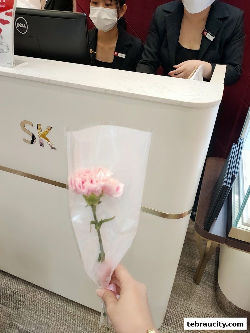 A hand holds a pink carnation in front of a jewelry store counter with staff.