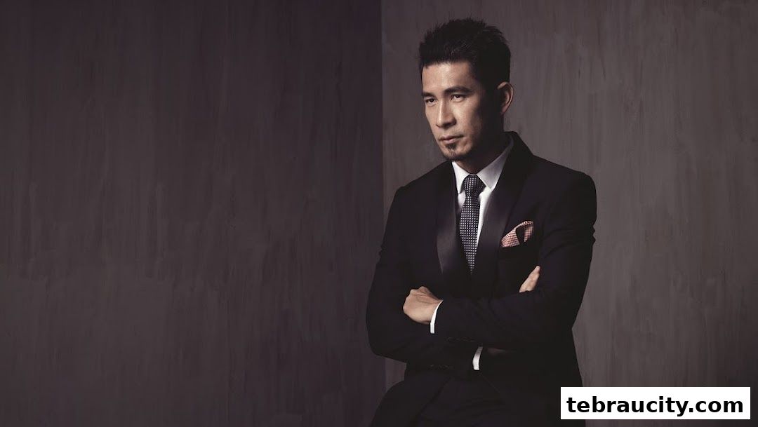 A man in a black suit with arms crossed against a textured gray background.