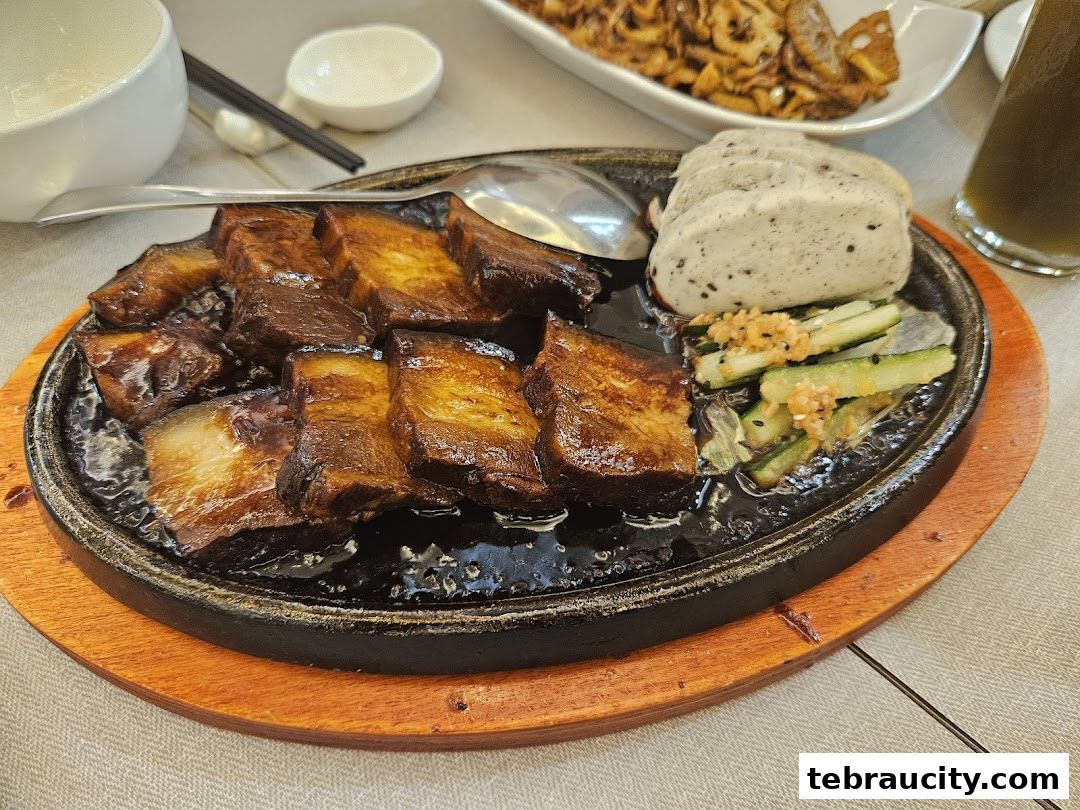 A sizzling plate of braised pork belly with a side of pickled cucumbers and steamed buns.