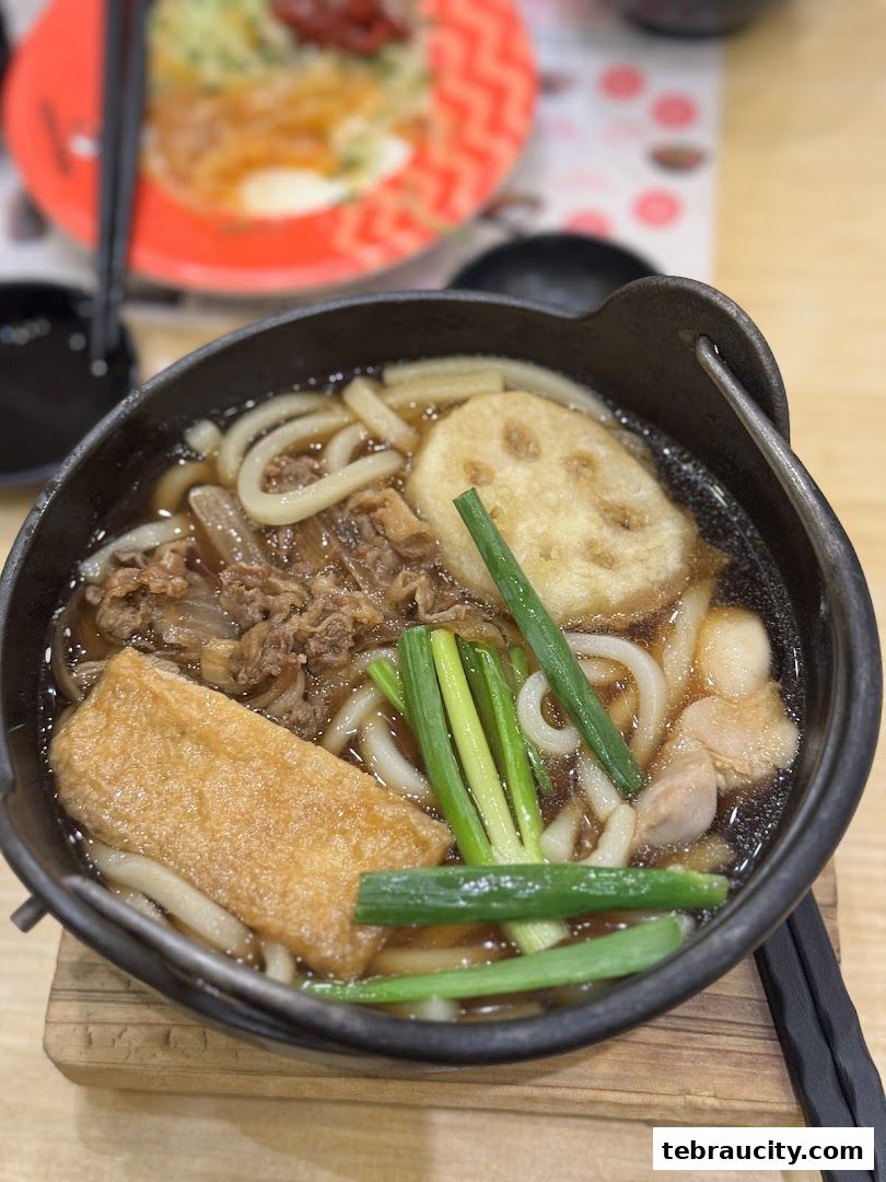 A close-up of a steaming bowl of udon noodles with beef, tofu, and lotus root.