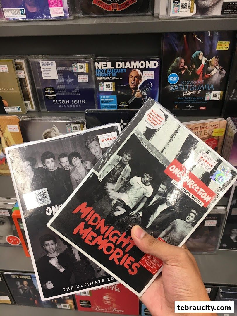 A hand holds two One Direction albums, 'Midnight Memories' and 'The Ultimate Edition', in front of a shelf of CDs and DVDs.