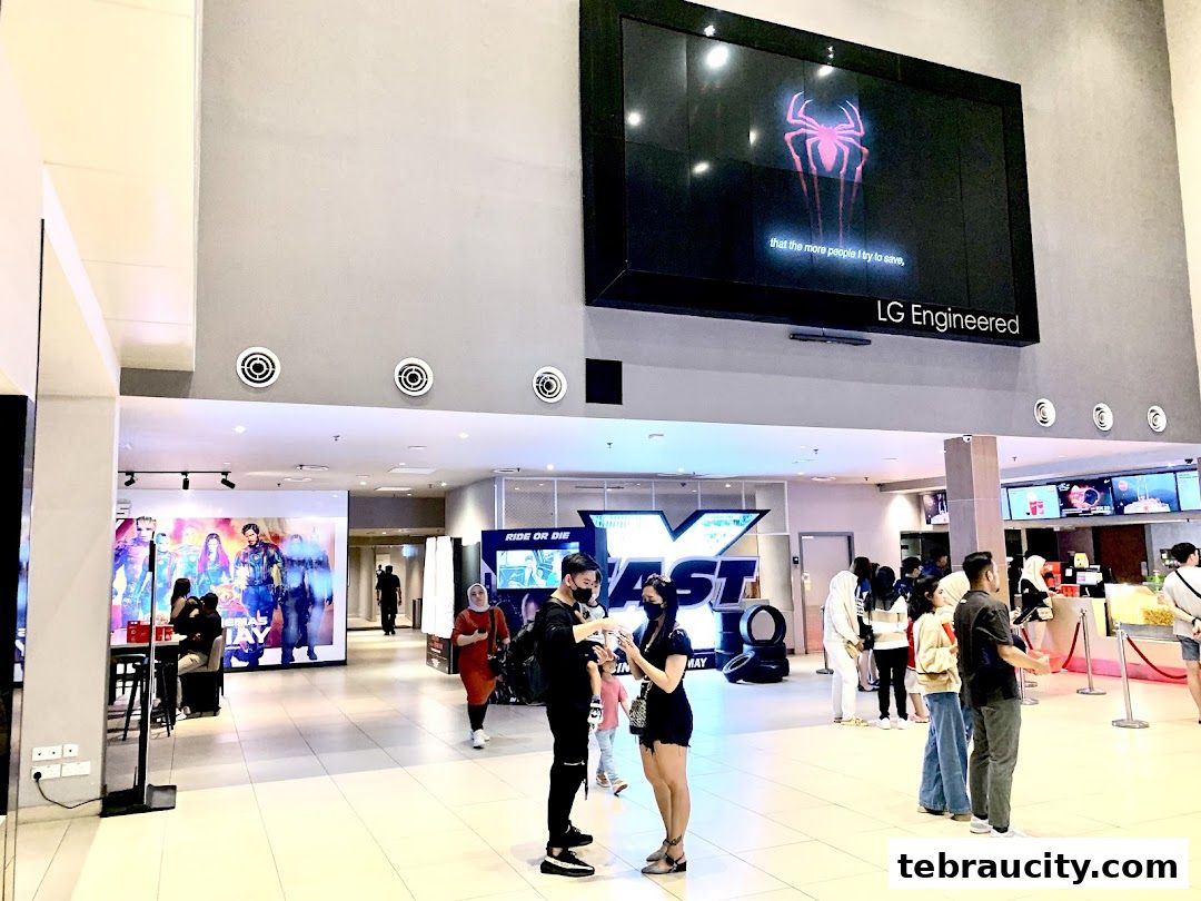 People queuing at a cinema lobby with movie posters and a large screen displaying a movie trailer.