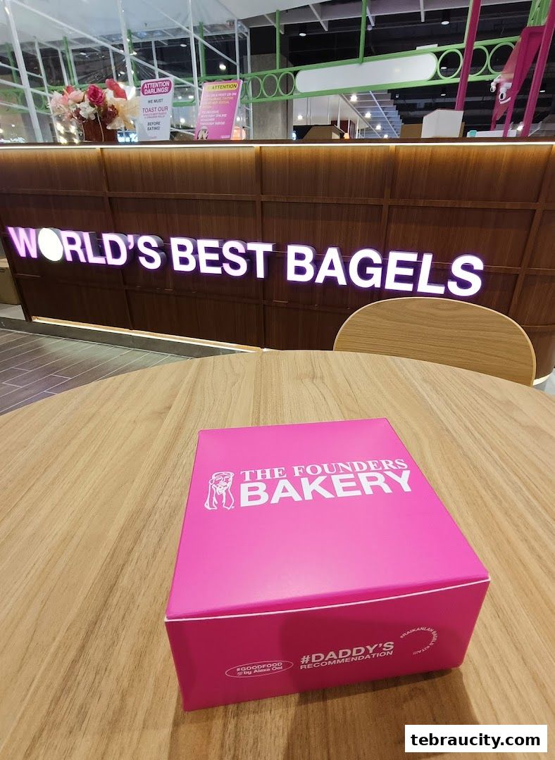 A pink box from The Founders Bakery sits on a wooden table in front of a counter with 'WORLD'S BEST BAGELS' signage.