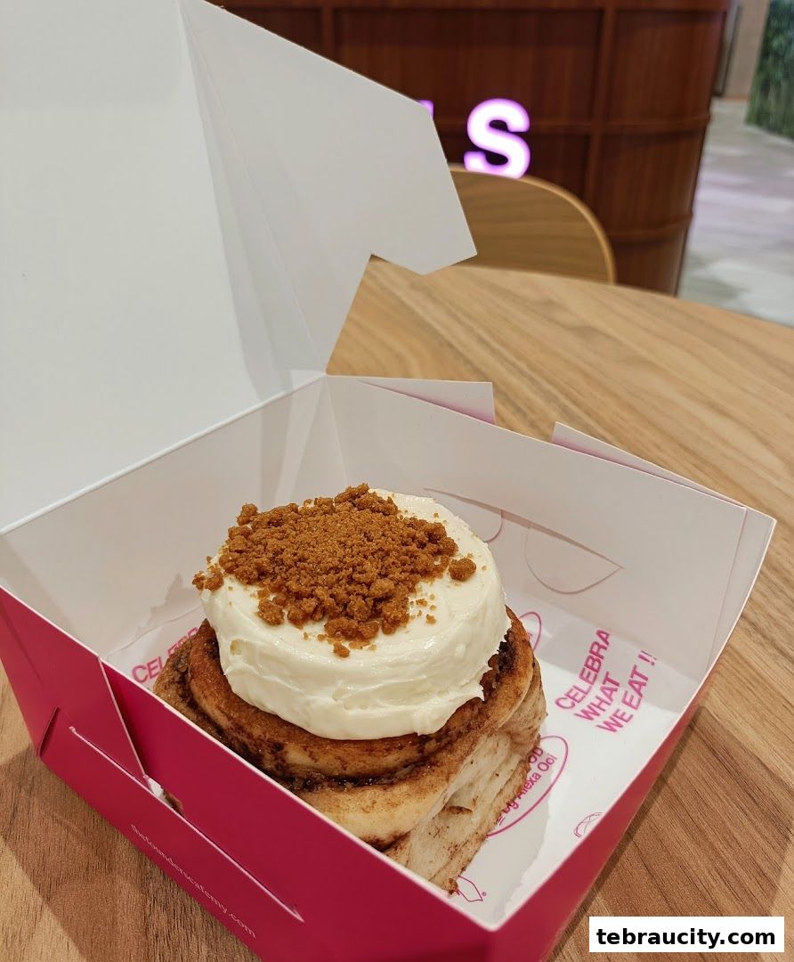 A delicious cinnamon roll with cream cheese frosting and cookie crumble topping in a pink box.