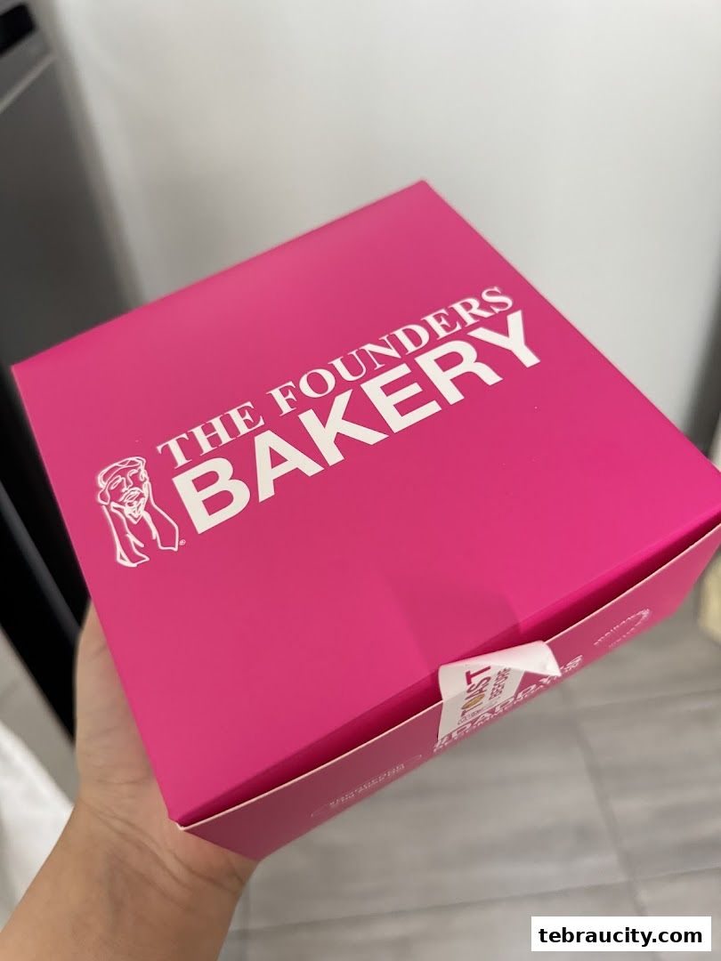 A hand holds a bright pink bakery box with the shop's logo.
