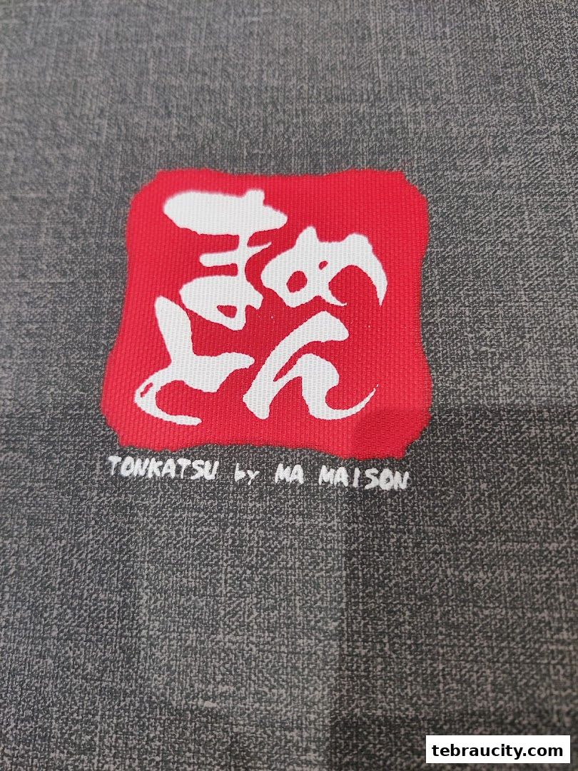 A close-up of the Tonkatsu by Ma Maison logo on a textured grey fabric.