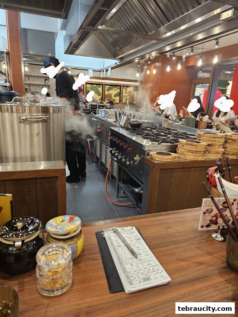 Busy ramen shop kitchen with chefs preparing food and customers dining.