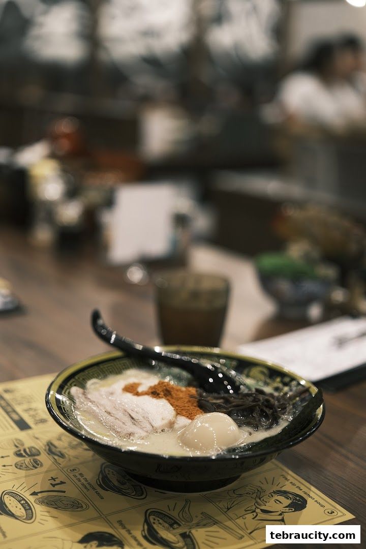 A steaming bowl of ramen with pork, egg, and spices sits on a table with a menu.