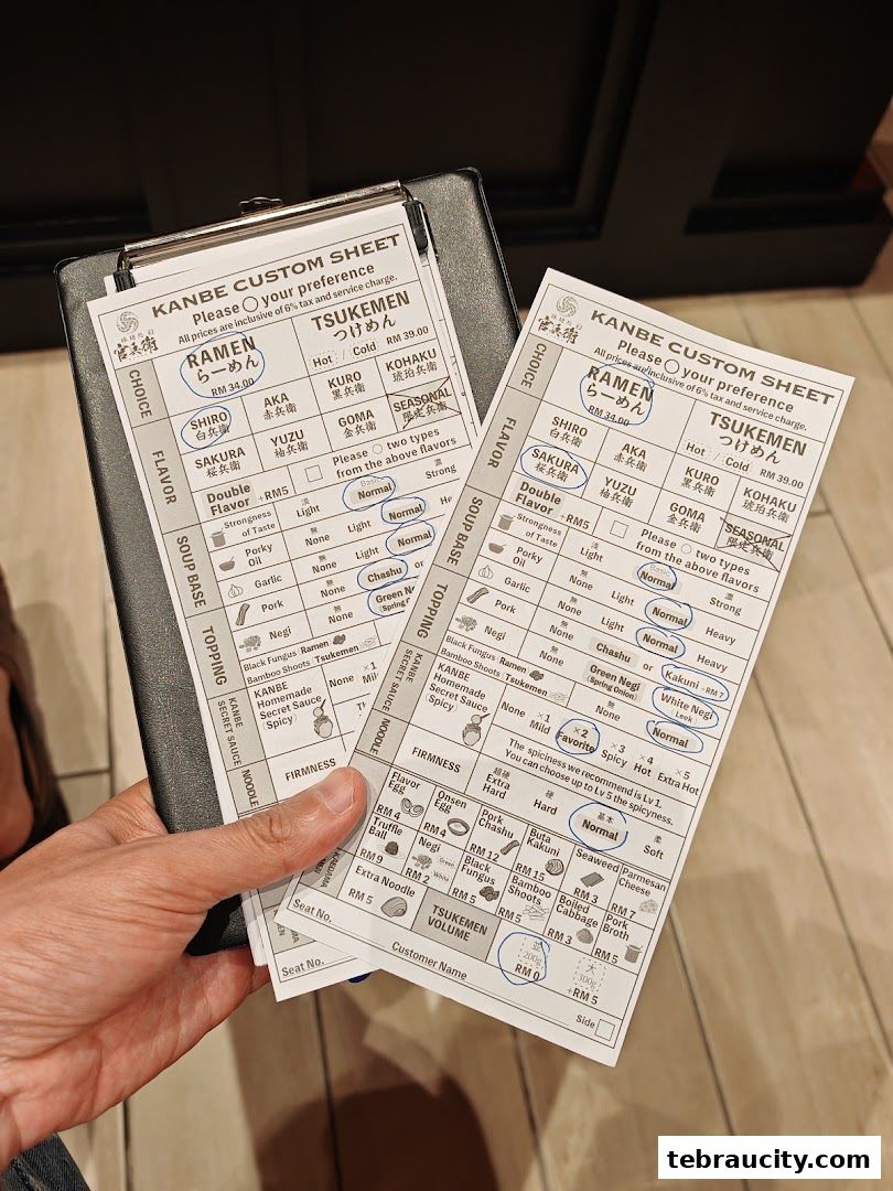 A person holding two custom order sheets for ramen and tsukemen at KANBE RAMEN JB.