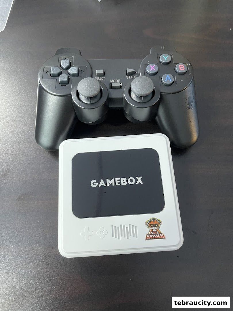 A black game controller rests on a dark surface next to a white Gamebox device.