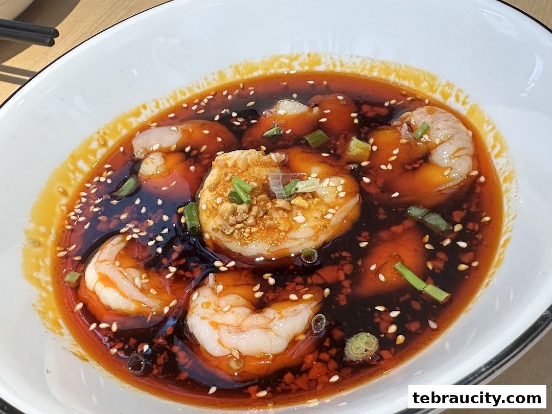 A close-up shot of a bowl of spicy shrimp dish with chili oil, sesame seeds, and chopped scallions.