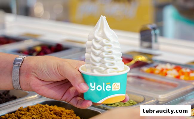A hand holds a cup of soft-serve frozen yogurt with various toppings in the background.