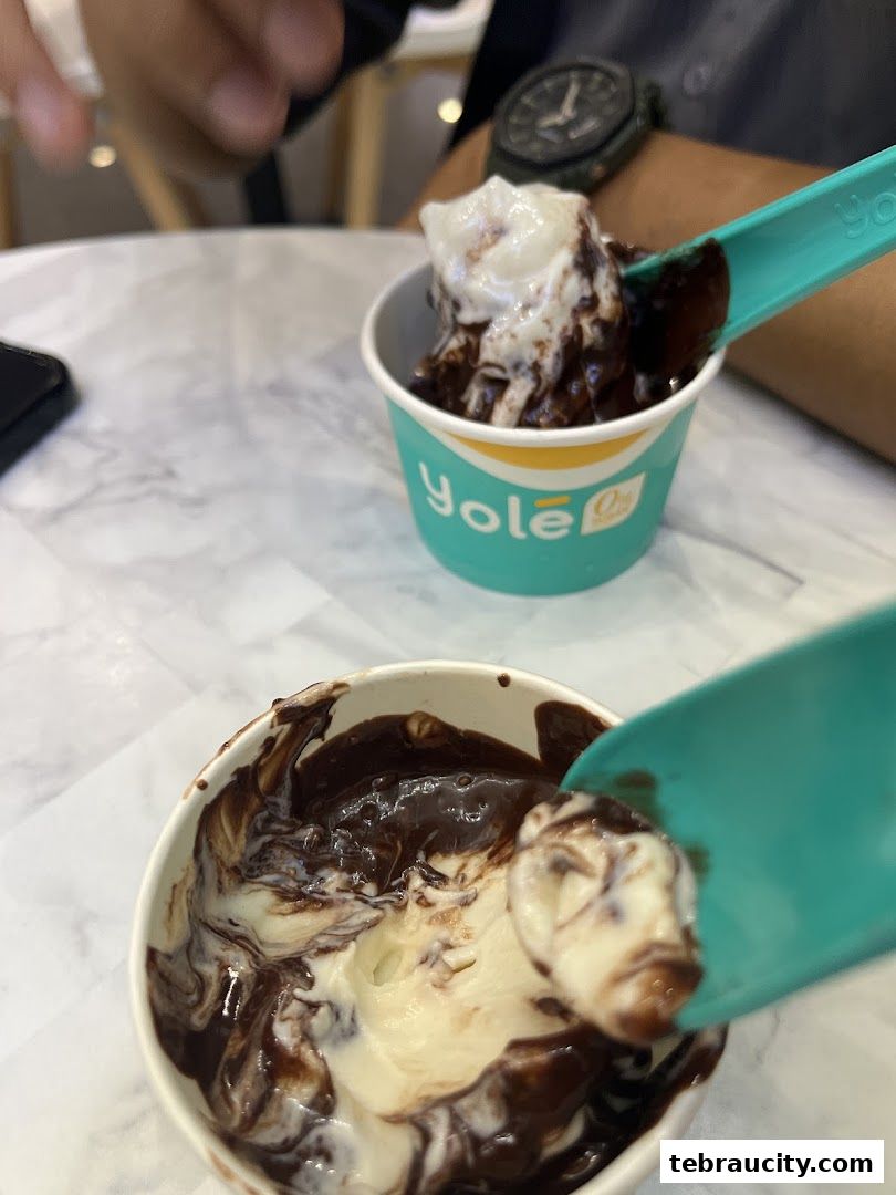 Two cups of frozen yogurt with chocolate sauce, being eaten with spoons.