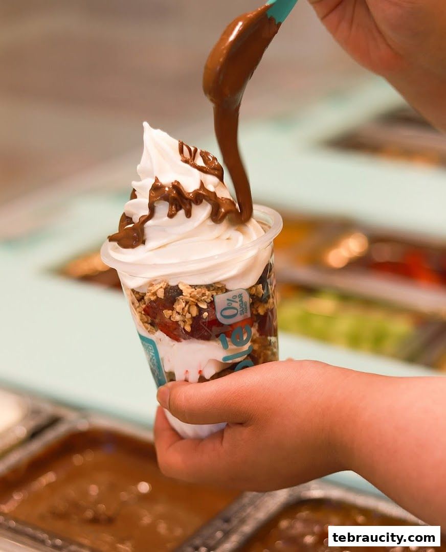 A hand holds a cup of frozen yogurt with toppings and chocolate sauce being drizzled on top.